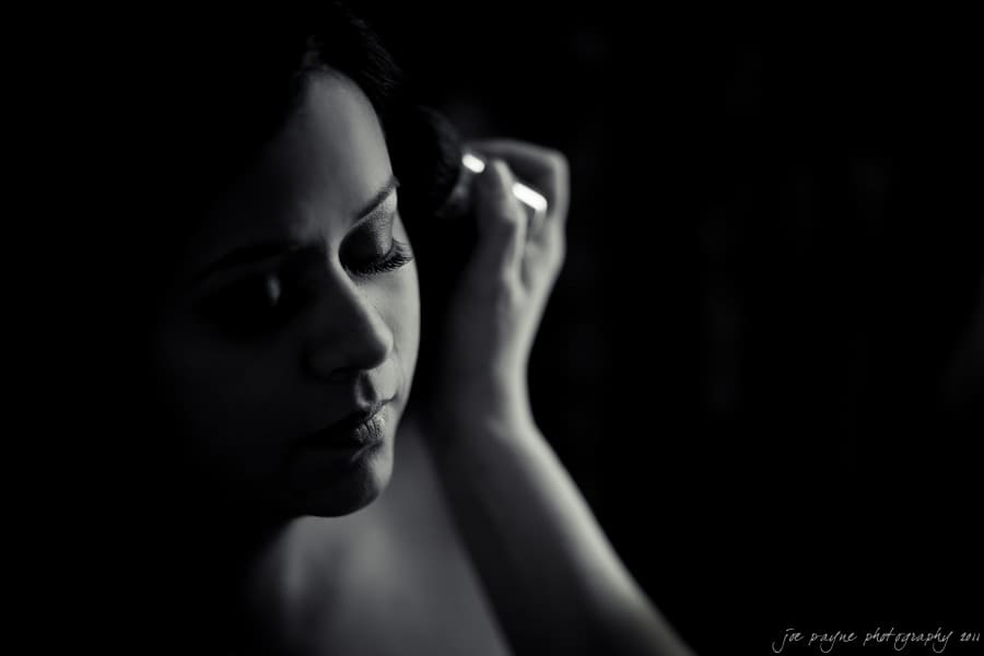 A black and white image of a person in low light, gently touching their hair, with a soft expression on their face. The lighting creates a dramatic contrast, highlighting their features against a dark background.