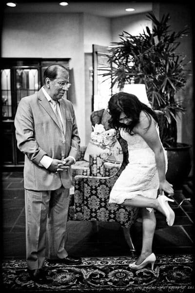 A bride in a white dress adjusts her high heel by a patterned chair, while an older man in a suit looks on with a small smile. They are indoors, with plants and furniture in the background. The image is in black and white.