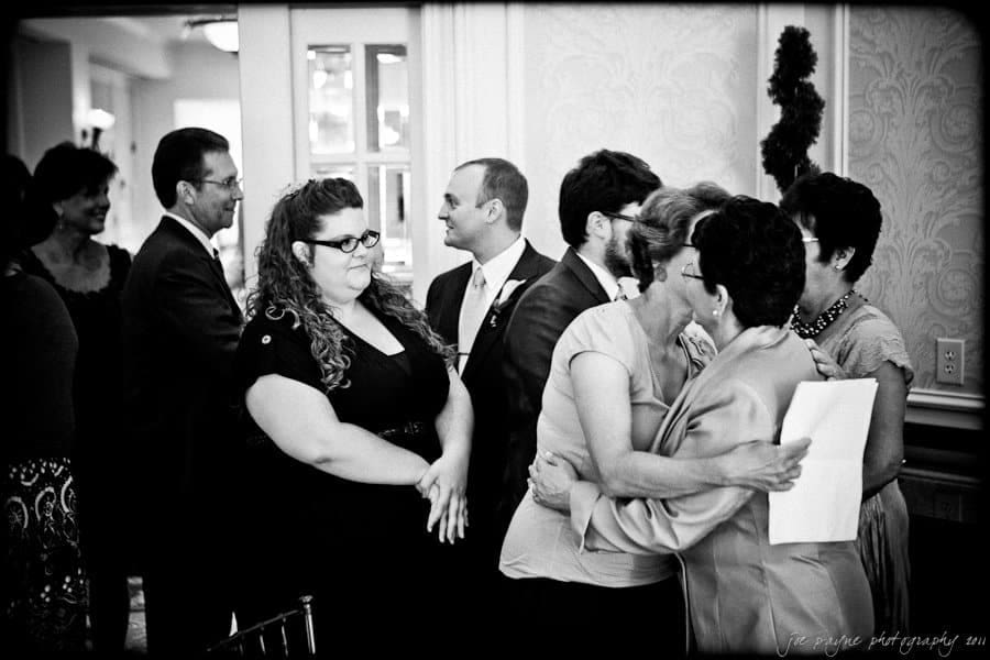 A black and white photo of a group of people having a conversation in what appears to be an indoor gathering. Some are dressed formally, and a woman is embracing someone while holding a piece of paper. Others are talking and observing.