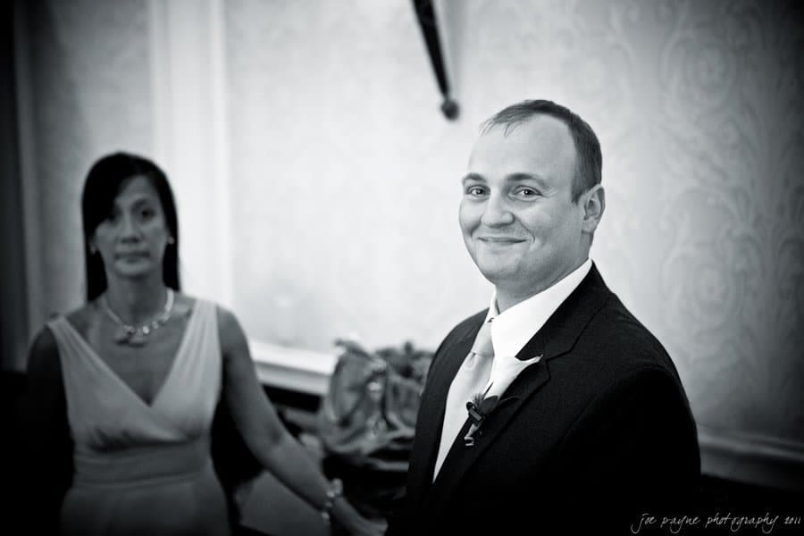 A smiling man in a suit stands in the foreground, while a woman in a dress with long hair stands slightly behind him. The setting appears to be indoors with elegant decor. The image is in black and white.