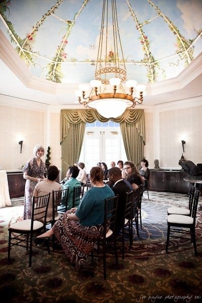 A small gathering of people seated in a formal room with elegant decor. A woman stands at the front, speaking. The ceiling features a painted sky and floral design. A chandelier hangs in the center, and large curtains frame the doorway.