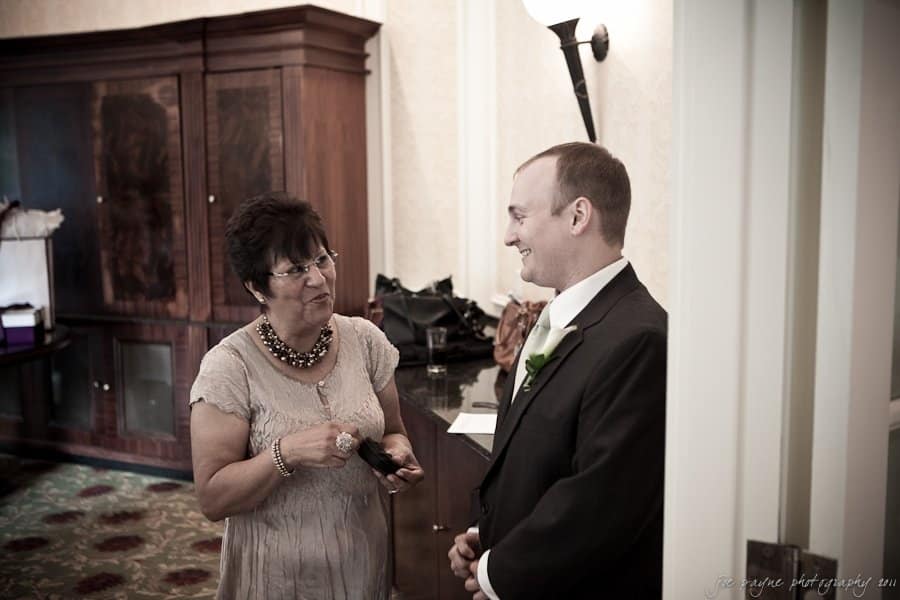 A woman and a man in formal attire stand indoors, smiling at each other. The woman wears a taupe dress and glasses, while the man is in a black suit with a boutonnière. A wooden cabinet and a lamp are in the background.