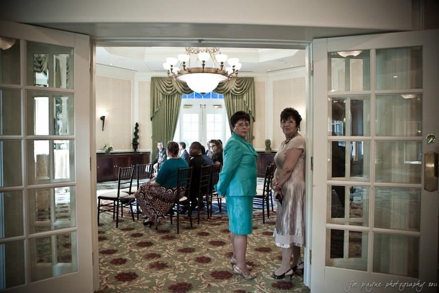 A group of people seated in a formal room with elegant decor, as seen from an open doorway. Two women in formal attire stand near the entrance, looking towards the camera. Chandeliers and large windows are visible in the background.