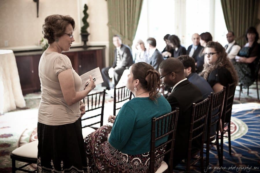 A woman stands and speaks to a seated group of people at an indoor event. The group is dressed formally, and they are seated in rows on chairs. The setting has a patterned rug and natural light coming from large windows.
