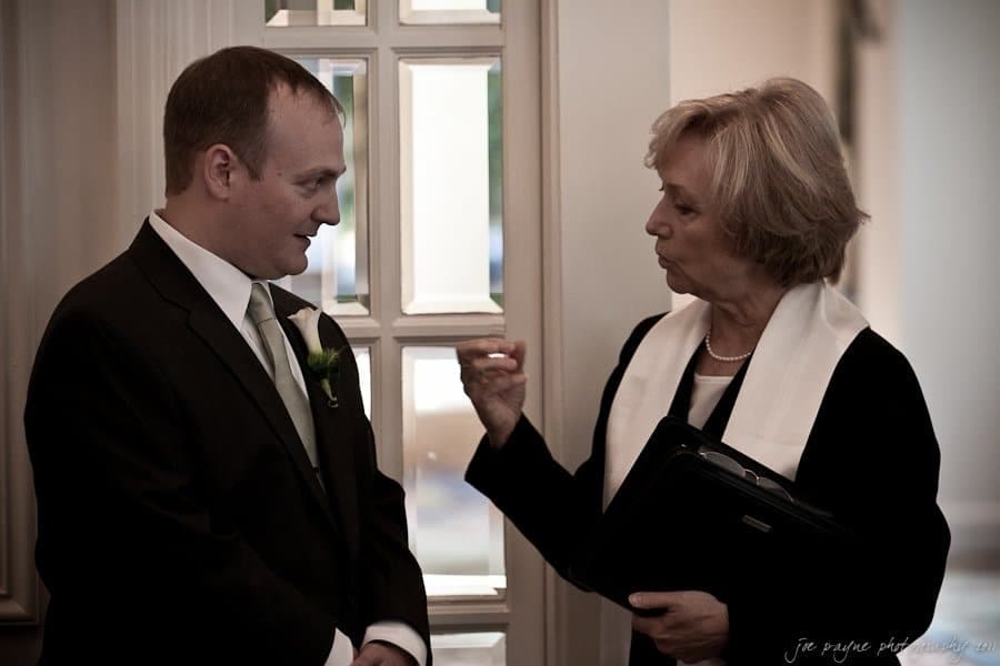 A man in a suit and tie is speaking with a woman wearing a white scarf and holding a folder. They are standing near a windowed door, appearing to have a discussion indoors.
