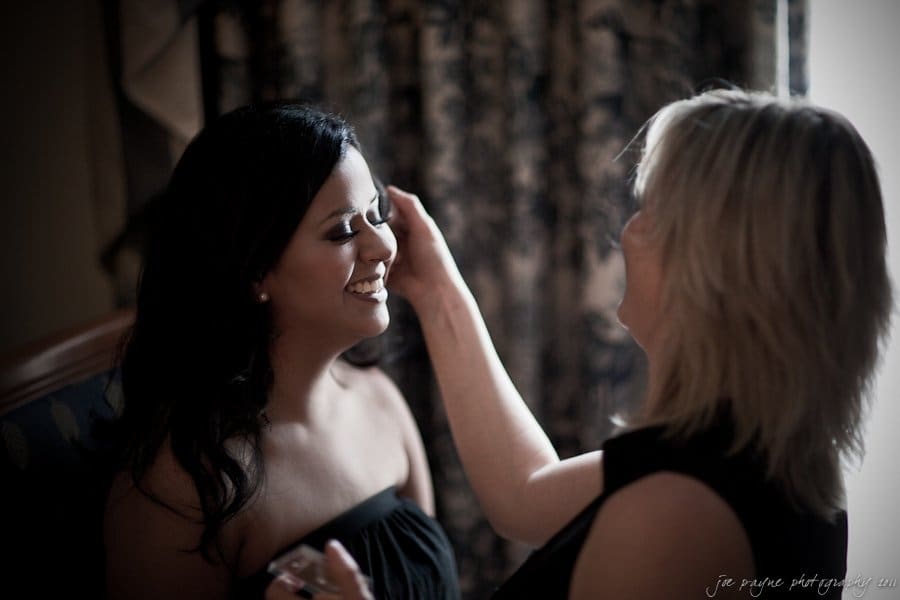 A woman with dark hair sits smiling, while another woman with blonde hair applies makeup to her face in a warmly lit room. The background features patterned curtains.