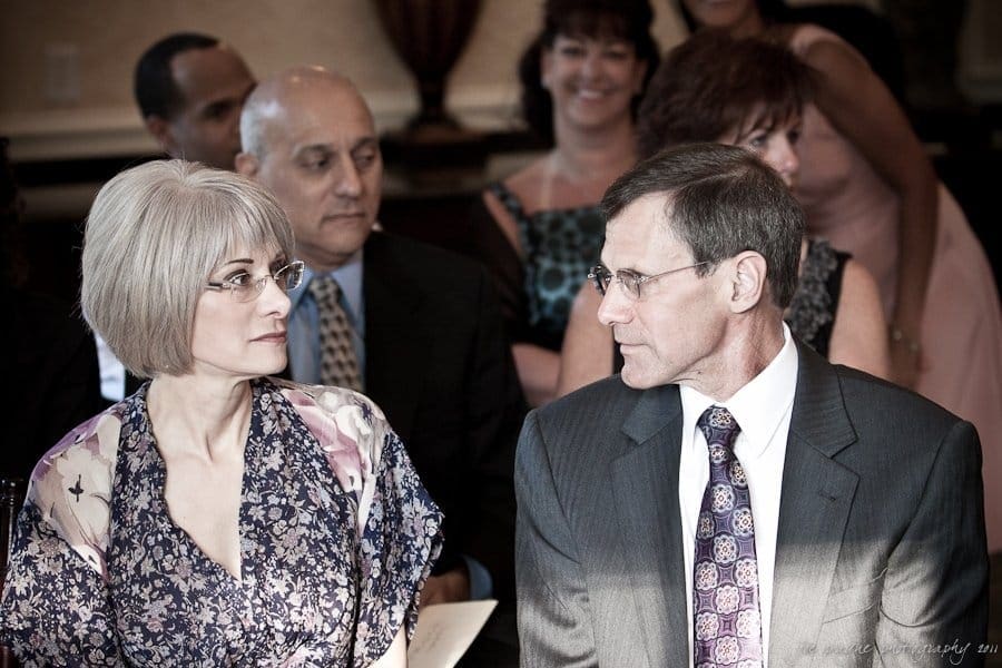 A man and woman in formal attire sit next to each other at an event, engaged in conversation. The man is wearing a suit and tie, while the woman wears glasses and a floral blouse. Several people are visible in the background, seated and smiling.