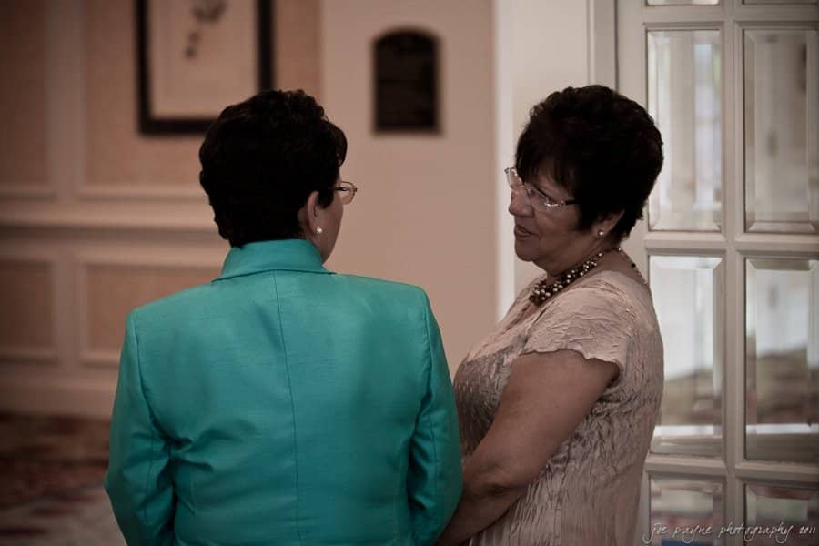 Two women stand facing each other in conversation. One wears a turquoise jacket, and the other is in a beige dress. They are indoors, near a window with white frames, in a room with neutral-colored walls and a patterned carpet.
