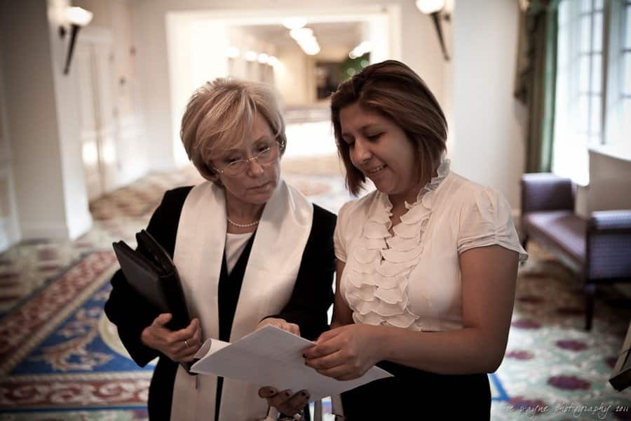 Two women in a well-lit hallway discuss a document. One wears glasses and holds books, while the other shows the paper. The space features patterned carpet and elegant lighting.