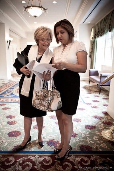 Two women stand in a well-lit room, engaged in conversation. One holds documents, while the other carries a bag and seems to be listening attentively. They are both dressed in professional attire, and the room features elegant decor.