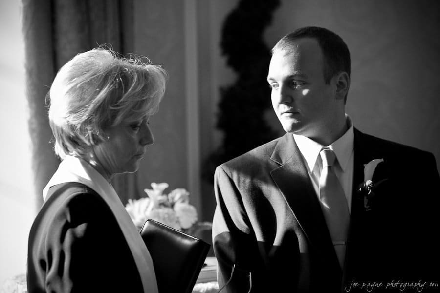 A black and white photo of a solemn indoor scene with two people dressed formally. A woman holds a book, while a man wearing a suit and tie stands nearby, both looking downward, suggesting a serious or reflective moment.