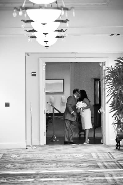 A black and white photo of a couple kissing in a hotel hallway. The man is in a suit, and the woman is in a dress holding a bouquet. The setting includes patterned carpet and decorative lighting.