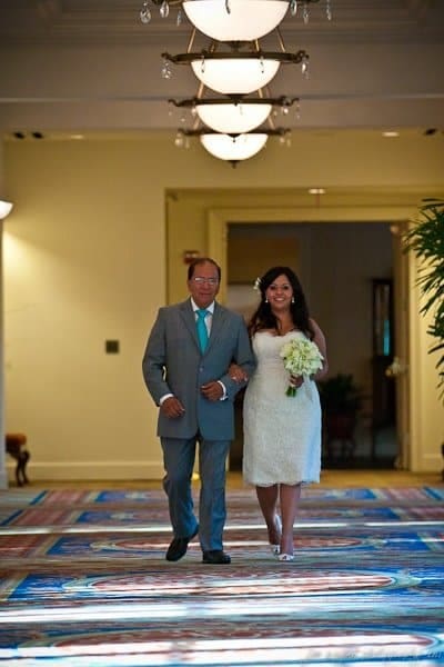 A bride in a white dress walks down an ornate, colorful carpeted aisle with an older gentleman in a gray suit. They are smiling, and she holds a bouquet of white flowers. The aisle is illuminated by overhead lights.