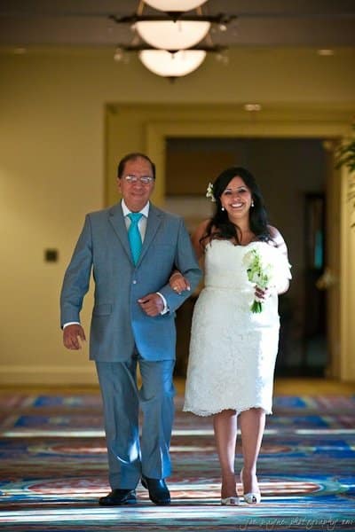 A bride in a white dress walks arm-in-arm with an older man in a gray suit and blue tie down a decorated aisle. She is smiling and holding a bouquet of white flowers. The setting appears to be indoors with soft lighting.