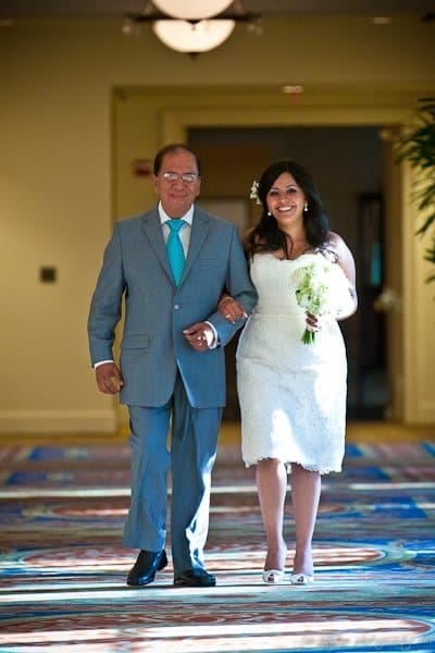 A bride in a knee-length white dress holds a bouquet and walks arm-in-arm with an older man in a gray suit and turquoise tie down a well-lit, decorated hallway.