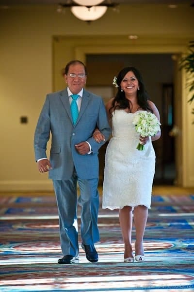 A bride in a white dress, holding a bouquet, walks arm in arm with a man in a gray suit and turquoise tie. They are smiling as they walk down an indoor aisle with colorful carpeting.