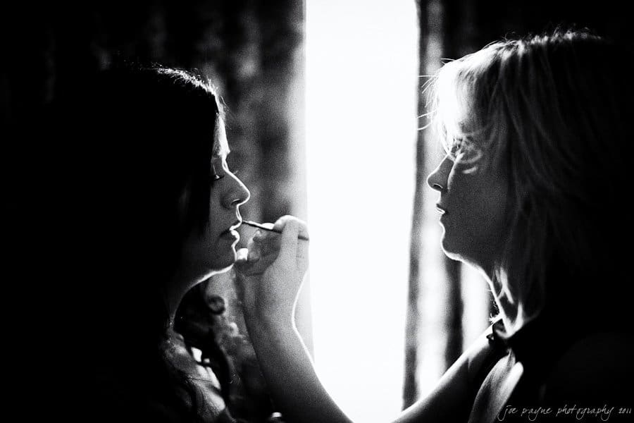 A black and white image shows a woman applying makeup to another womans lips. They are facing each other, illuminated by soft light from a window in the background. Curtains are partially visible.