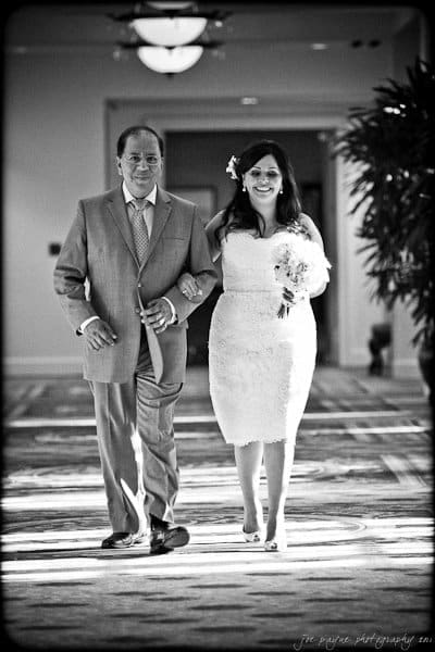 A man in a suit walks down an indoor aisle with a woman in a white lace dress holding a bouquet. Both are smiling, and there are potted plants visible in the background. The image is in black and white.