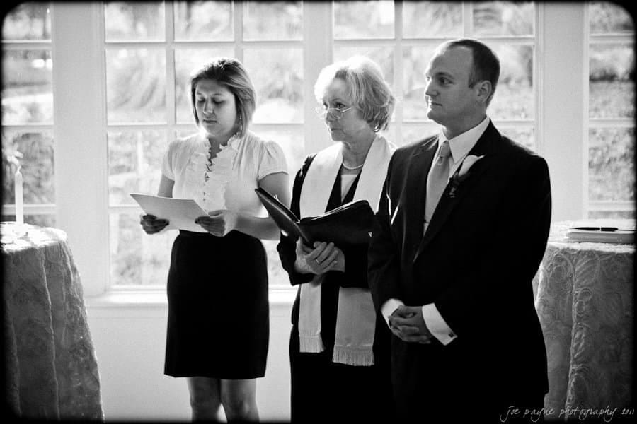 Three people stand indoors during a ceremony. A woman on the left is reading from papers, a middle-aged woman in the center holds a book, and a man in a suit on the right looks ahead. They are in front of a window with natural light.