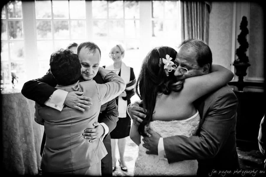 A black and white photo shows two couples embracing warmly inside a room with large windows. The expressions suggest a joyful and emotional moment. Elegant curtains frame the windows in the background.