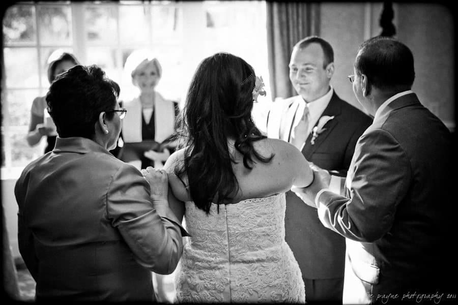 A black-and-white photo of a wedding ceremony. A bride in a strapless dress and groom in a suit face each other, holding hands. Two people stand behind the bride, and one person is visible in the background. Sunlight streams through a window.
