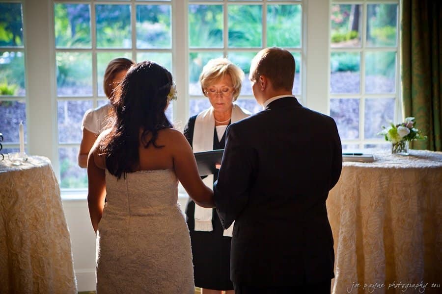 A couple stands before a woman officiating a wedding ceremony in a sunlit room. The bride wears a white dress, and the groom is in a dark suit. They face the officiant, who holds a book, with two witnesses nearby.
