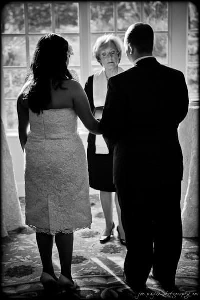 A black and white photo of a couple facing each other, holding hands, in front of an officiant during a wedding ceremony. Natural light streams in through a large window behind them.