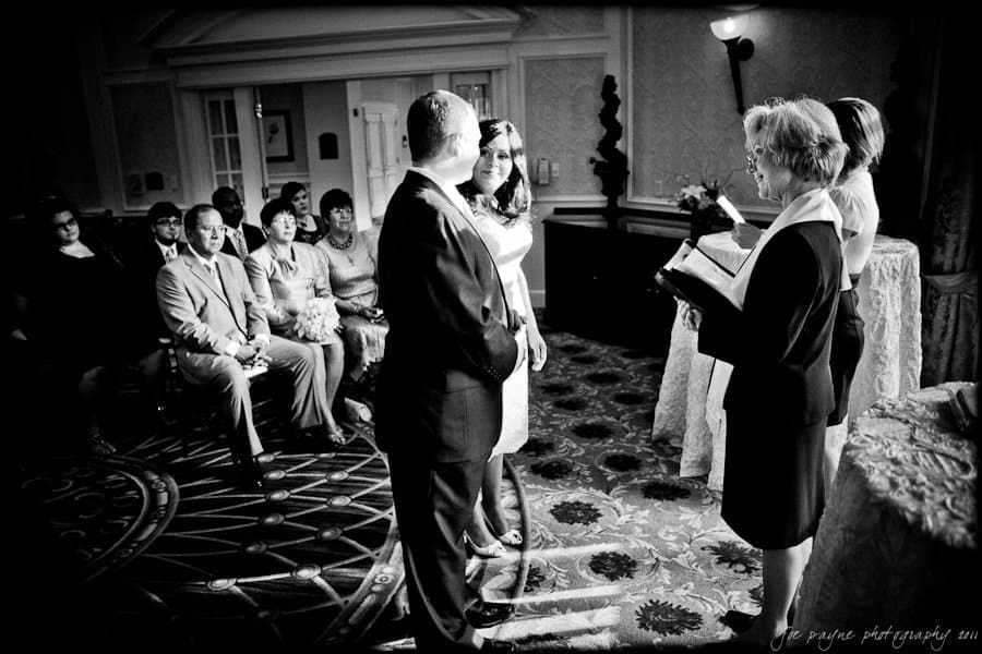 A black and white photo of a couple standing together during a wedding ceremony. The officiant is reading from a book, and guests are seated, watching the couple. The setting is an ornate room with decorative carpeting and elegant wall designs.