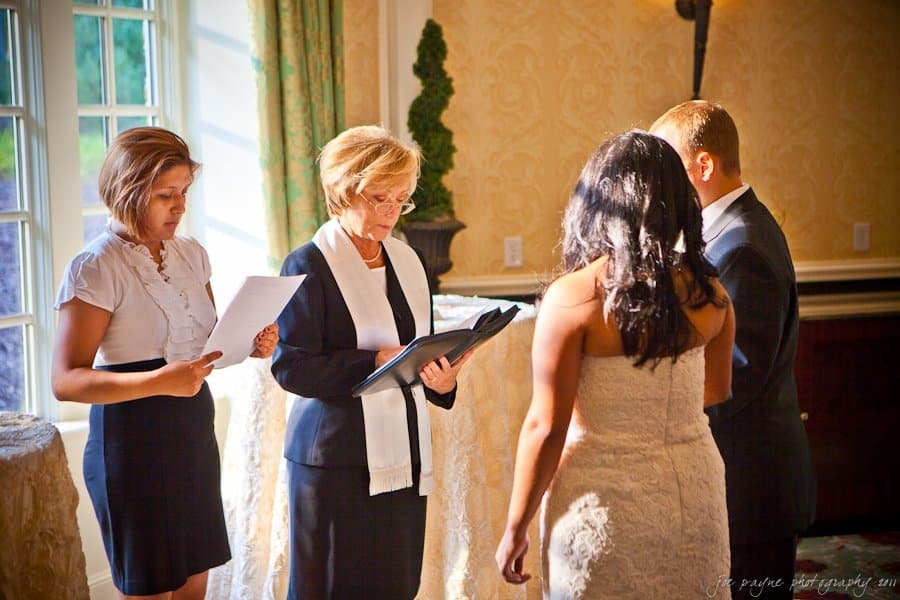 A wedding ceremony with a couple standing before an officiant. The bride is in a lace gown, and the groom is in a suit. A witness or assistant stands nearby holding papers. Sunlight streams in through a window, creating a warm, intimate atmosphere.