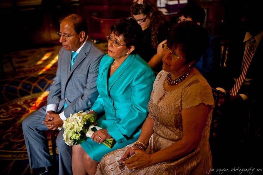 Three individuals seated indoors, two women and one man. The woman in the center holds a bouquet, wearing a teal suit, while the others are in formal attire. The setting appears to be a formal event with patterned carpet and ambient lighting.
