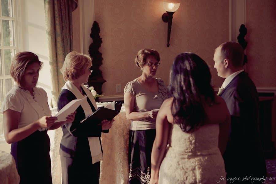 A group of five people standing in a warmly lit room, participating in a ceremony. Two women are holding open books, one woman appears to be speaking, and a couple stands together facing them. A window with light filtering in is in the background.