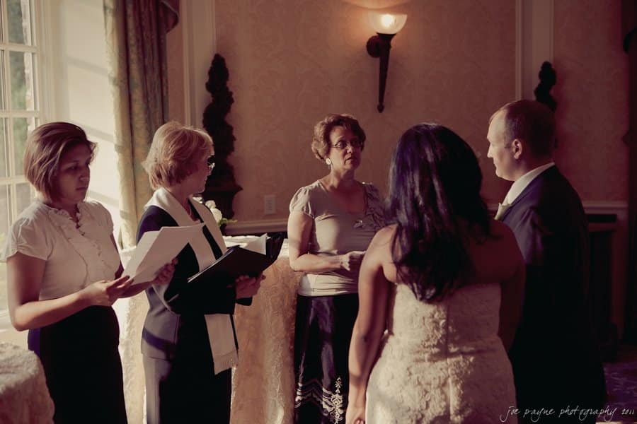 A wedding ceremony with four women officiating, two holding books. A couple stands facing them, with the woman in a white dress and the man in a suit. The room is warmly lit with ornate wallpaper and a large window.