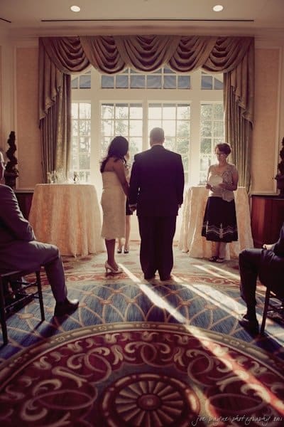 A couple stands in front of a sunlight-drenched window during a ceremony, with a woman holding papers nearby. The scene is set in an elegant room with decorative curtains and a patterned rug. Guests are seated, observing the event.
