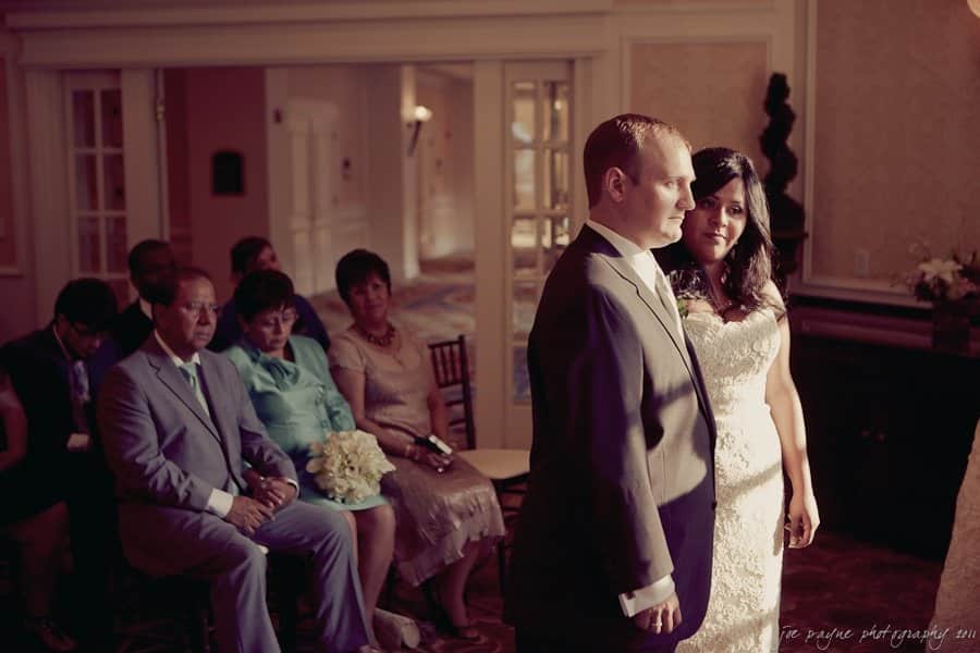 A couple stands together at the front of a dimly lit room, with seated guests watching them. The bride wears a white gown, and the groom is in a dark suit. The atmosphere is intimate and formal.