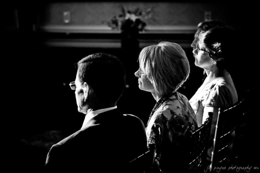A black and white photo of three adults seated in a dimly lit room. The light highlights their profiles as they face forward. There are chairs visible, and a flower arrangement is blurred in the background.