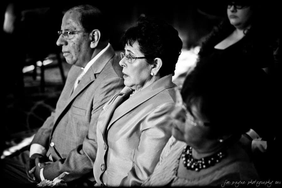A black and white photo of three older adults seated in a row, dressed formally. The focus is on the woman in the middle, wearing a light-colored suit. They appear attentive, possibly attending a ceremony or event.