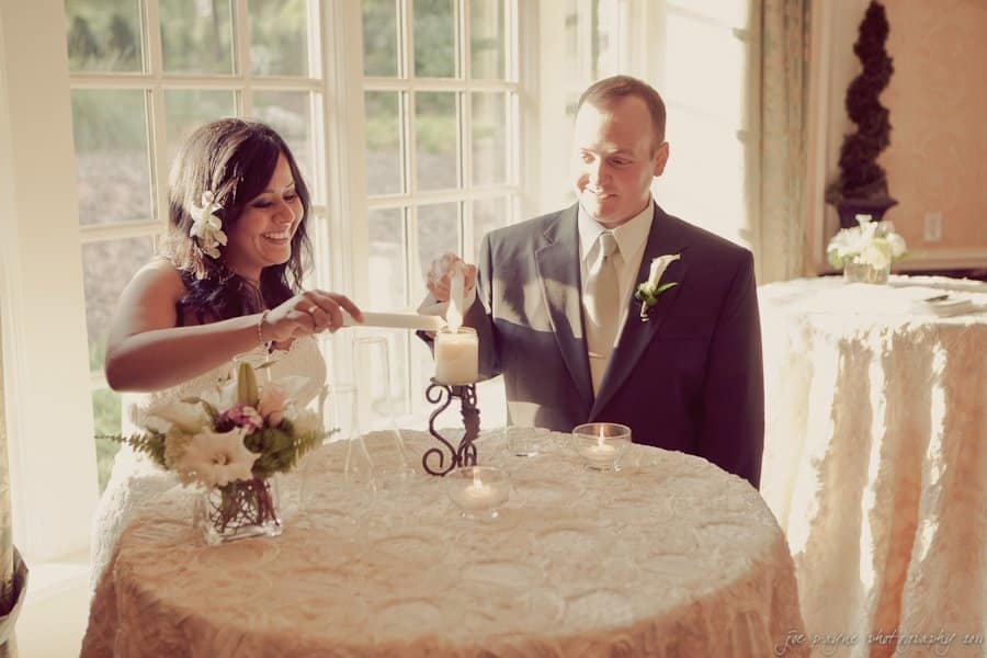 A woman and a man, dressed formally, light a large candle together at a round table draped with a lace tablecloth. The scene is warmly lit by natural light streaming through a large window in the background. A floral arrangement sits on the table.