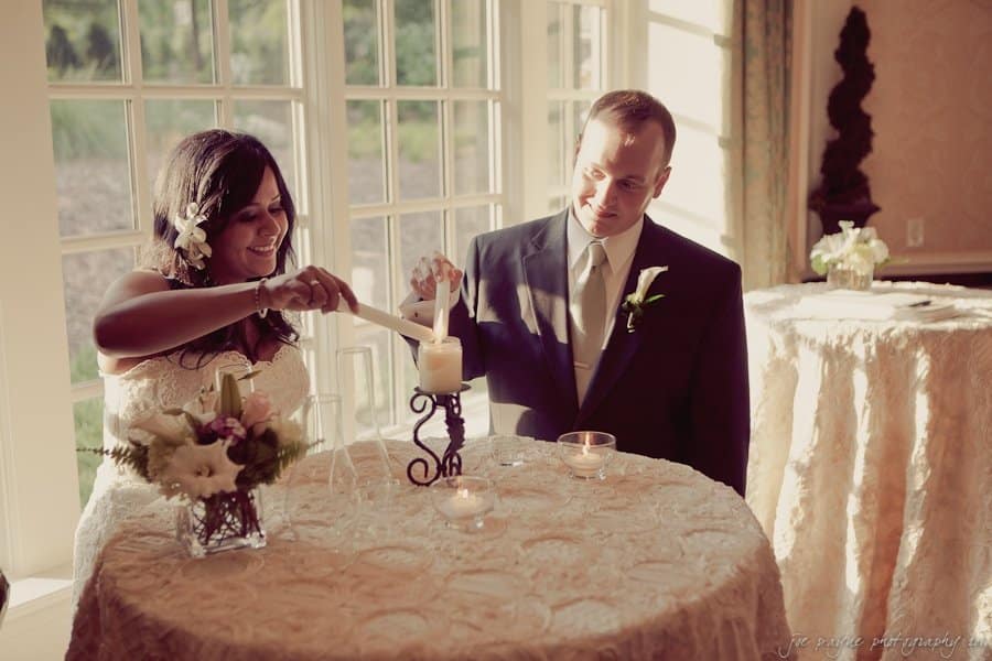A bride and groom are lighting a unity candle together during a wedding ceremony. They are standing at a round table with decorative cloth and floral arrangements, near large windows letting in sunlight.