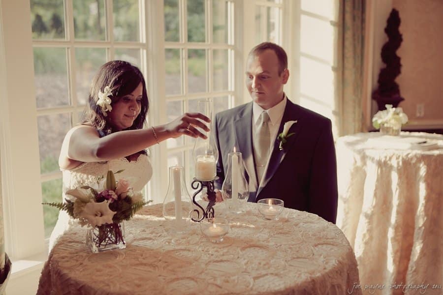 A bride and groom participate in a candle lighting ceremony during their wedding. The bride, in a white dress, adjusts a candle, while the groom, in a suit, looks on. Theyre standing by a window with decorated tables around them.