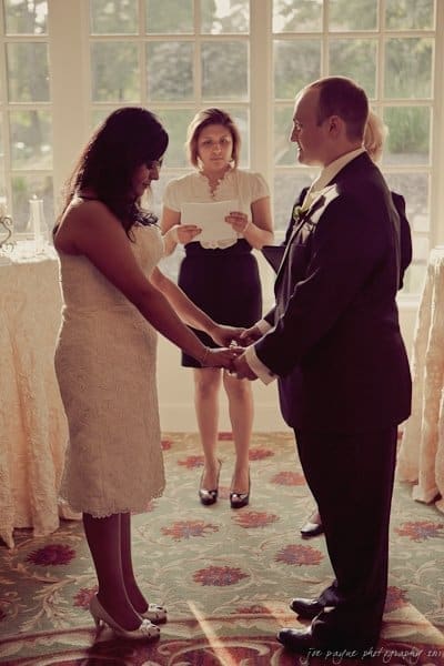 A couple stands facing each other, holding hands in a sunlit room, as an officiant reads from a paper in the background. The scene suggests a wedding ceremony. Light filters through a large window behind them.