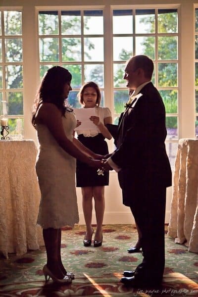 A couple stands holding hands during a wedding ceremony, with a woman officiating. They are backlit by sunlight streaming through large windows, creating a romantic atmosphere. The scene is indoors with decorative tables in the background.