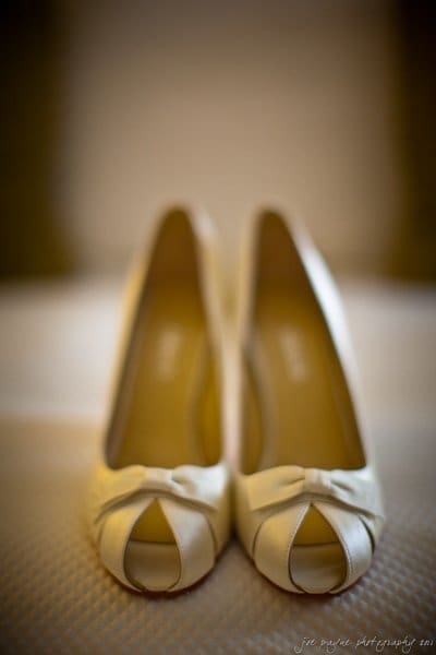 A close-up of a pair of elegant, cream-colored peep-toe heels with decorative bows on a soft, textured surface. The shoes are aligned side by side, creating a symmetrical and stylish composition.