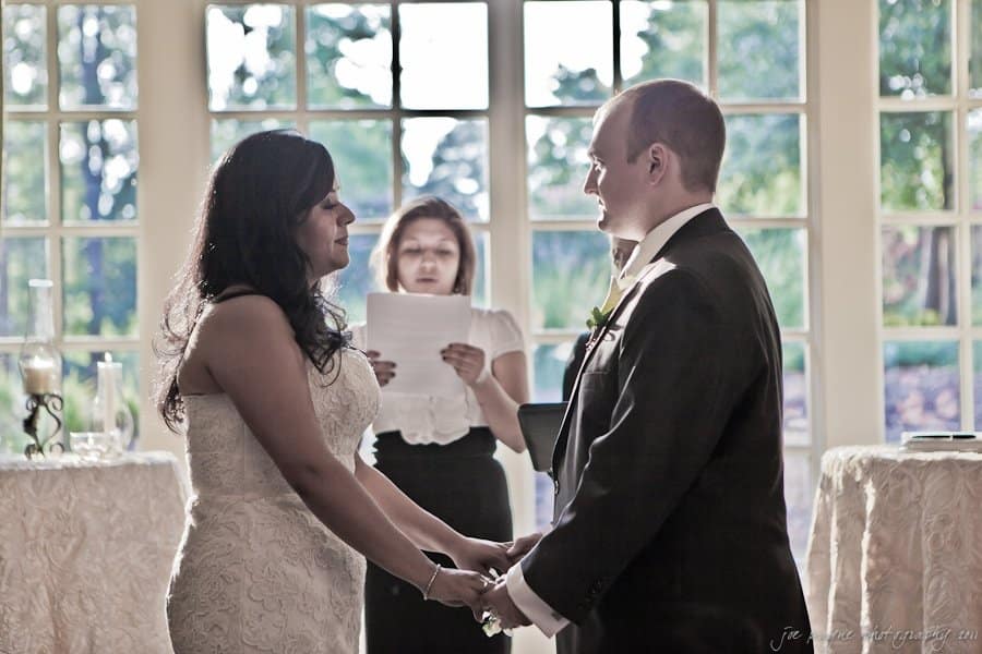 A couple stands holding hands facing each other during a wedding ceremony. A woman in the background reads from a paper. Soft light filters through large windows, creating a warm ambiance.