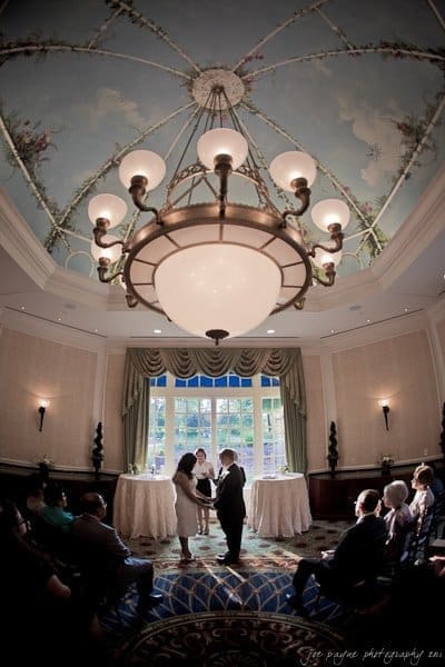 A couple stands at the altar in an elegantly decorated room with a large chandelier and painted ceiling. Guests are seated, observing the ceremony. Natural light streams through the tall windows in the background.