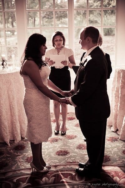 A bride and groom hold hands during a wedding ceremony. The bride is wearing a white dress and the groom is in a suit. An officiant stands behind them, reading from a paper. They are in front of large windows. The setting is softly lit.