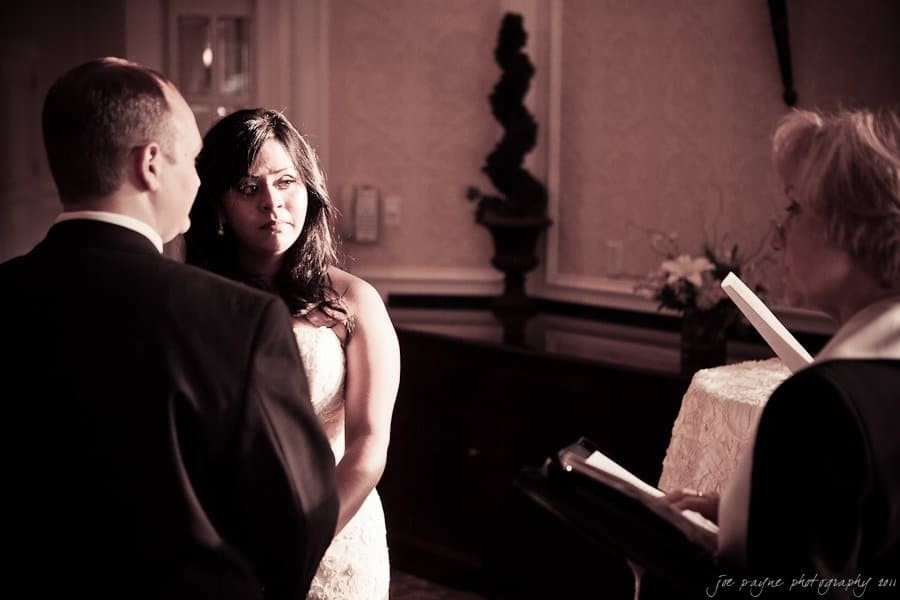 A bride and groom stand facing each other during a wedding ceremony in a softly lit room. The bride looks attentively at the groom, while an officiant stands nearby with an open book.