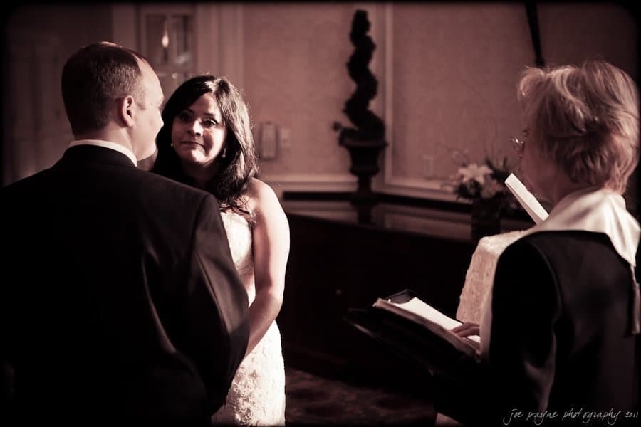 A bride and groom stand facing each other during a wedding ceremony. The bride gazes at the groom while an officiant stands to the side holding a book. The setting is softly lit with elegant decor.