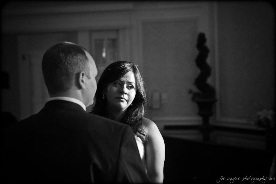 A woman in a white dress gazes at a man in a suit. The dimly lit, elegant room has decorative molding and a potted plant. Shadows and light create a dramatic effect.