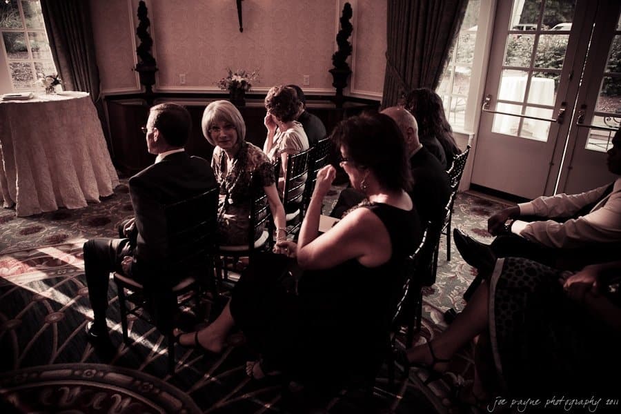 A group of people seated inside, looking towards a semi-lit area. Curtains frame the windows on the right, and sunlight streams in through them, casting soft shadows on the floor. The ambiance is warm and intimate, suggesting a formal gathering.