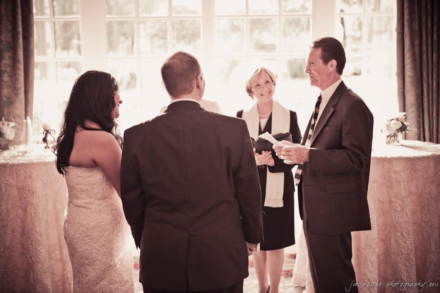 A wedding ceremony with a bride in a lace dress and a groom facing a smiling officiant. A woman stands beside the officiant holding a book. Soft lighting and elegant decor create a warm atmosphere.